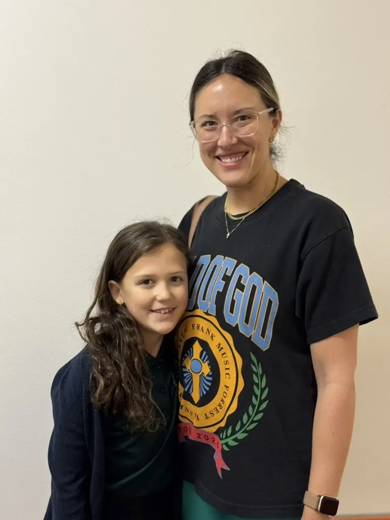 A woman and a young girl standing close together indoors, both smiling at the camera.