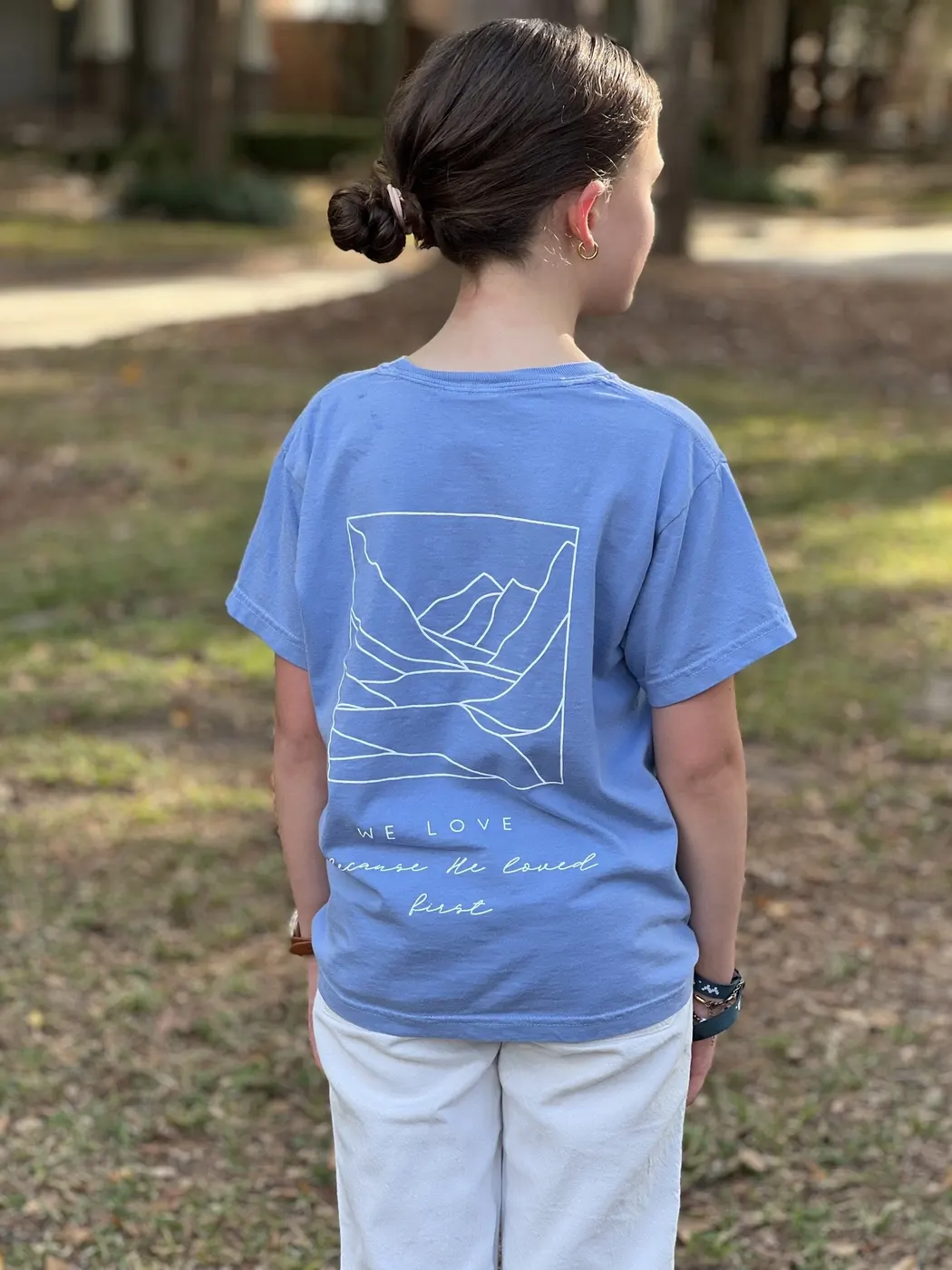 Youth tee in soft blue, with white mountain line art and the words We Love because He loved first