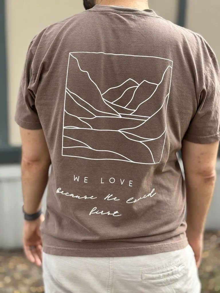 Adult tee in earth brown, with white mountain line art and the words We Love because He loved first