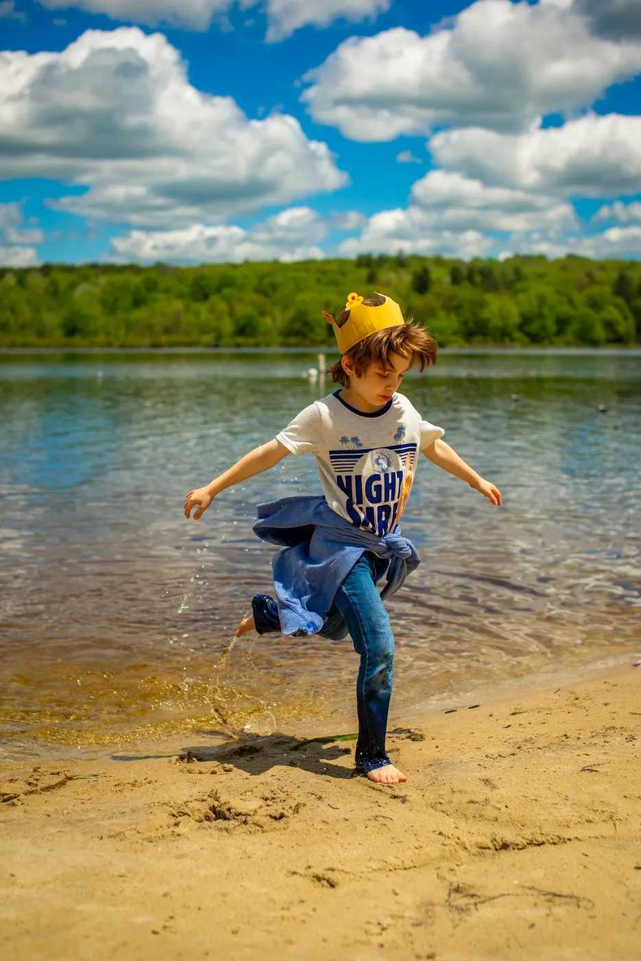 A boy in a paper crown kicks up a splash of water at the edge of a lake, with green trees and a blue sky in the background.